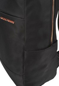 Mochila de tela negra con una textura suave, que cuenta con un cierre prominente y un bolsillo lateral. El logotipo "SKECHERS" está grabado en naranja.