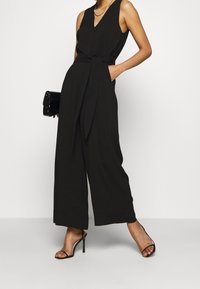 Černý bezrukávový overal s výstřihem do V, širokými nohavicemi, látkovým pásem v pase a bočními kapsami. V kombinaci s černými sandály na vysokém podpatku a psaníčkem.