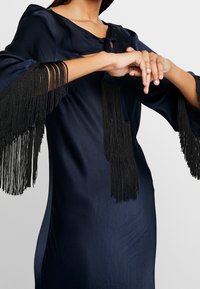 Vestido de satén azul marino con un ajuste relajado. Presenta detalles de flecos negros en las mangas y un escote redondeado con cierre de lazo.