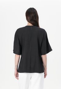 Top noir à manches courtes en tissu léger avec un col rond et une couture arrière droite, porté avec un pantalon blanc.