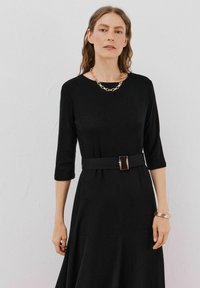 Czarna sukienka z rękawami typu trzy czwarte, z wcięciem w talii, wyposażona w pasek z prostokątną klamrą, gładka faktura i zwiewny krój. Złota łańcuszkowa naszyjnik.