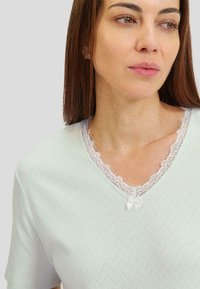 Top de manga corta verde menta con escote en V, que presenta un borde de encaje blanco y un pequeño detalle de cinta. Patrón en zigzag texturizado en toda la prenda.