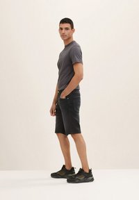 T-shirt gris, shorts en denim noir et baskets camouflées noires. Le modèle se tient de profil, les mains dans les poches, avec un fond neutre.