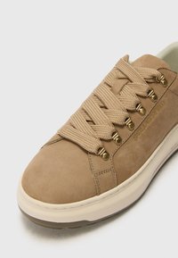 Beige suede sneaker with thick woven laces, gold-tone eyelets, and a low-profile rubber sole; minimalistic design with a clean finish.