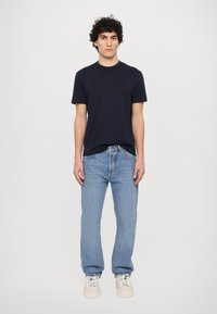 Σκούρο μπλε βαμβακερό t-shirt με ανάγλυφο λογότυπο, συνδυασμένο με φωτεινά μπλε τζην. Το μοντέλο φοράει λευκά Sneakers, και στέκεται μπροστά σε έναν ουδέτερο φόντο.