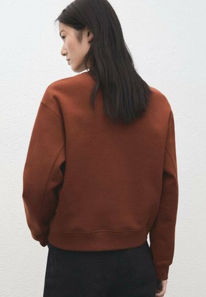 Sudadera color óxido con un ajuste relajado, puños y dobladillo acanalados, y textura suave. Se lleva con jeans negros, vista desde atrás.