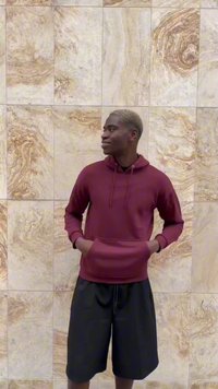Bordeaux hoodie met kangoeroezak, gedragen met zwarte shorts; staat voor een gestructureerde beige en gouden muur.