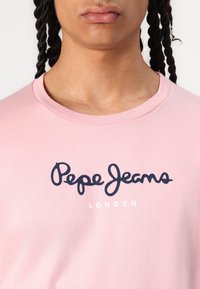 Rožinės spalvos medvilnės marškinėliai su tamsiai mėlynu "Pepe Jeans" logotipu ir baltais "LONDON" užrašais, apvaliu kaklu ir trumpomis rankovėmis.