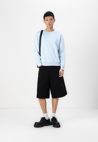 Light blue crewneck sweatshirt, black knee-length shorts, and chunky black shoes. Model stands with a black shoulder bag, on a white background.