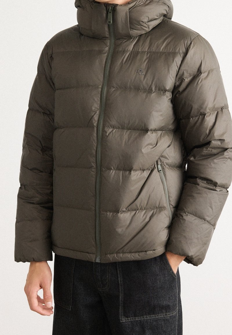 Homme portant une doudoune olive à fermeture éclair avec col montant et les mains dans les poches d'un pantalon en denim bleu foncé.