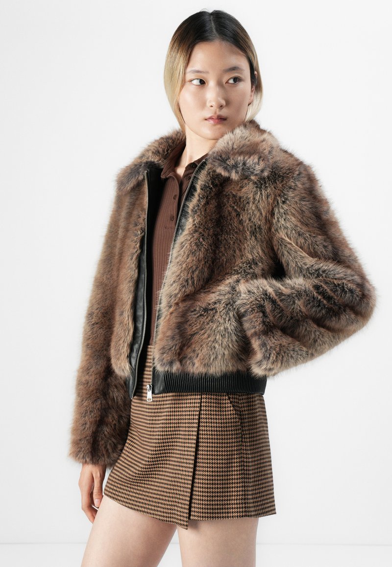 Kobieta w brązowej kurtce z futra syntetycznego, na ciemno-brązowej bluzce i mini spódnicy w brązową kratę, patrząca w bok.