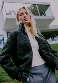 Temno siva flis jakna z visokim ovratnikom in zadrgo spredaj, z teksturirano tkanino in rebrastimi manšetami, oblečena čez belo majico.