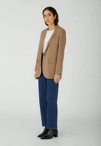 Young woman standing, wearing a beige blazer, white top, blue jeans, and black ankle boots against a plain light background.