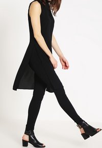 Svart ärmlös tunika med sidofickor, tillsammans med svarta leggings och öppna klacksandaler med ankelremmar och blockklackar.