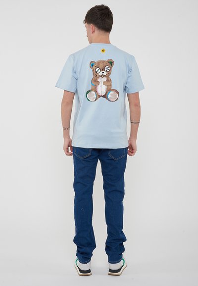 T-shirt azzurro chiaro in cotone, con una grafica di un orso sulla schiena in marrone, dettagli in bottone neri e accenti colorati. Indossa jeans scuri.