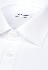 Gros plan sur le col et les boutons d'une chemise blanche, montrant l'étiquette de la marque "seidensticker" à l'intérieur du col.