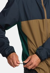 Větruodolná bunda s barevnými bloky v námořnické modré, zelené a béžové barvě. Lehký materiál s nastavitelnou šňůrkou a elastickými manžetami.
