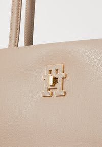 Sac à main texturé beige avec un fermoir en or proéminent mettant en avant des formes géométriques et un petit accent rectangulaire. Deux poignées robustes.