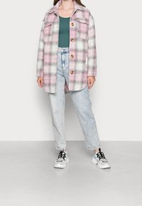 Karierte Jacke in Pink, Grau und Weiß mit einem Kragen und Brusttaschen; braune Knöpfe; getragen über einem grünen Oberteil und kombiniert mit hellblauen Jeans.
