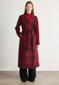 Palton din lână cu carouri roșii, cu o curea, guler tip rever, două buzunare laterale și o siluetă lungă, asortat cu pantaloni negri cu croială largă.