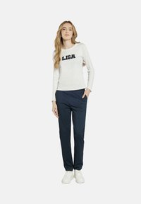 Biała koszula z długim rękawem z napisem "LISA" w granatowym kolorze, w zestawie z granatowymi spodniami w prążki. Model ma na sobie białe trampki, stoi na jednolitym tle.