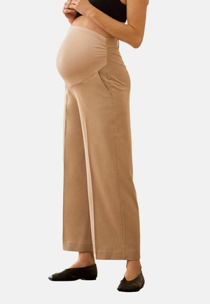 Brune graviditetsbukser med høj talje, sidelommer, glat tekstur og løs pasform, kombineret med sorte flade sko.