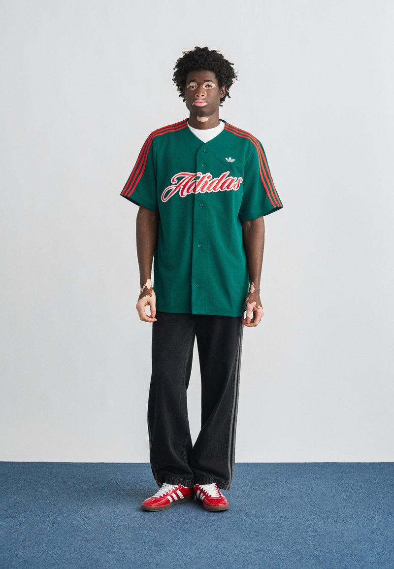 Maillot vert à boutons avec manches à rayures rouges et logo "Adidas", associé à un pantalon large noir et des baskets rouges à rayures blanches.