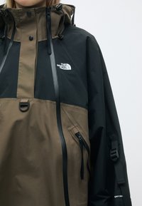 Brūna un melna ūdensnecaurlaidīga jaka ar kapuci, izceļoties ar rāvējslēdzējiem, regulējamiem sašaurinājumiem un logotipu uz krūtīm. Strukturēta auduma.