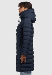 Marinblå pufferjacka med hög krage och justerbar huva. Har en quiltad design, sidofickor och ribbade cuffs för värme.