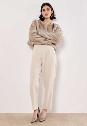 Kurz geschnittene Jacke aus Kunstfell in hellem Taupe, maßgeschneiderte cremefarbene Hose mit Falten und glänzende taupefarbene High Heels, die vor einem neutralen Hintergrund steht.