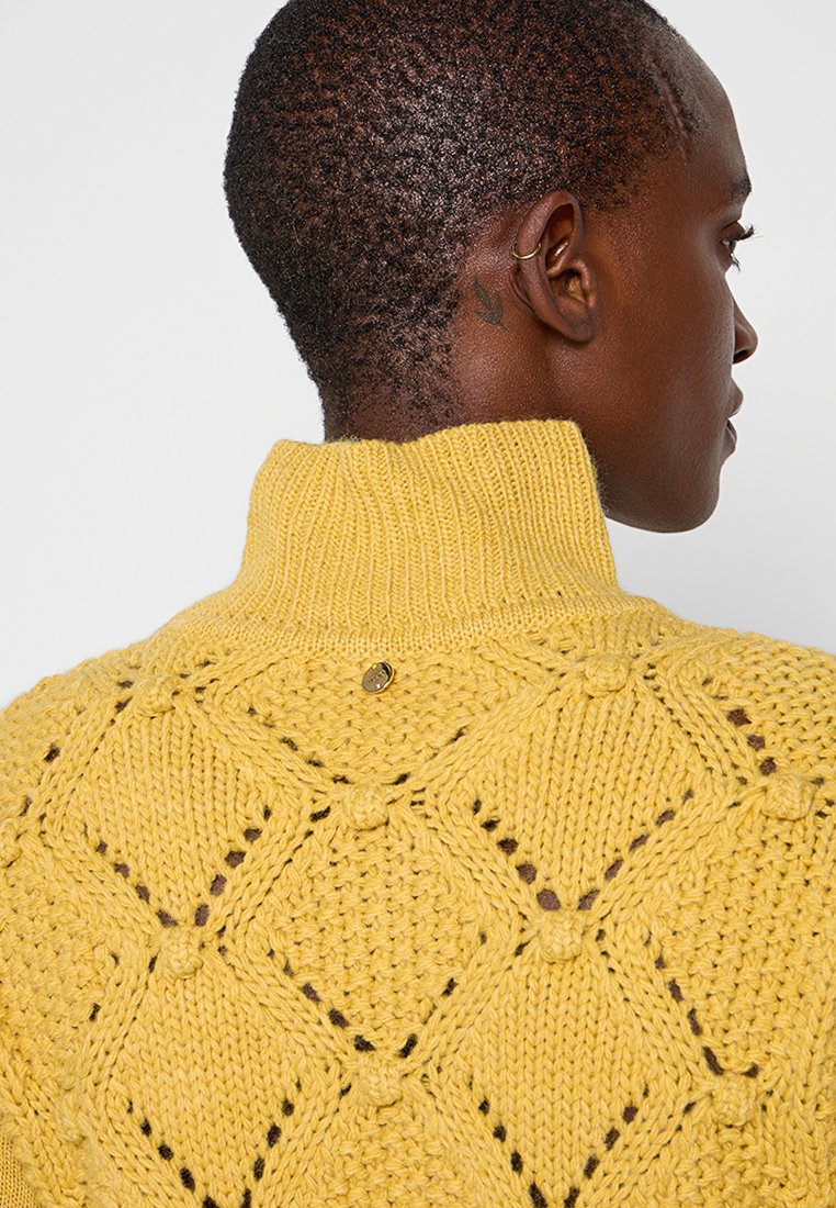 Maglione giallo lavorato a maglia con colletto alto, caratterizzato da motivi a diamante e dettagli tessurizzati sul retro, accentuato da un piccolo bottone in metallo.