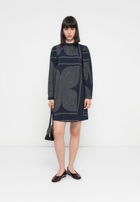 Abito della marina con maniche lunghe, caratterizzato da motivi geometrici bianchi. Vestibilità ampia, scollatura tonda, abbinato a scarpe basse nere e una borsa piccola.