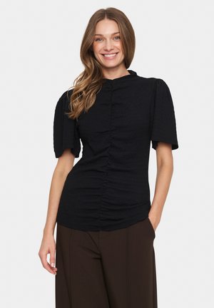 Glunderende vrouw met lichtbruin haar draagt een gerimpeld zwart shirt met korte mouwen en donkerbruine broek, poseert met één hand in de zak op een witte achtergrond.