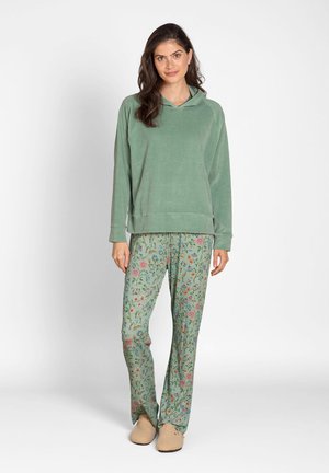 Woman standing wearing a green hoodie, green floral patterned pants, and beige slippers against a plain light background.