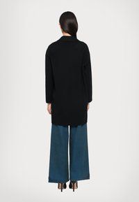 Schwarzer, übergroßer Mantel mit Kragen, langen Ärmeln und offenem Rücken, kombiniert mit weit geschnittenen blauen Jeans und schwarzen High Heels.