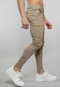 Pantalons skinny beiges avec poches avant et design ajusté, associés à des baskets blanches. Texture lisse et couleur unie. Vue latérale entièrement visible.