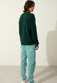 Grüner gestrickter Pullover mit rundem Ausschnitt, kombiniert mit hellblauen gestreiften Cargo-Hosen. Das Model steht leicht seitlich, um das Outfit zu präsentieren.