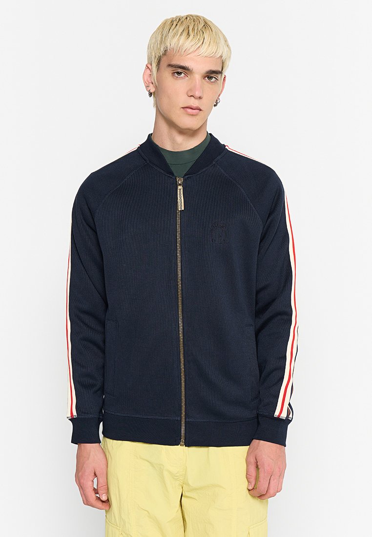 Marineblaue Zip-Jacke aus geripptem Material mit rot-weißen Streifenakzenten an den Ärmeln. Verfügt über einen Stehkragen und einen Reißverschluss.