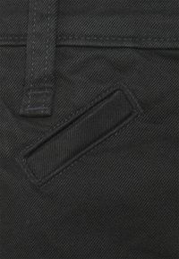 Black denim fabric with prominent diagonal ribbing and a rectangular slit pocket. Visible stitching along edges for added detail.
