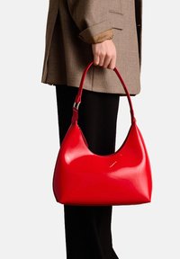 Red patent leather handbag with a curved shape and a single shoulder strap. Smooth texture with a glossy finish, held in a hand.
