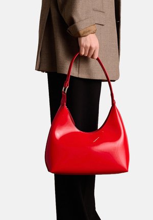 Rode leren handtas van patenteren met een gebogen vorm en een enkele schouderriem. Soepele textuur met een glanzende afwerking, vastgehouden in een hand.