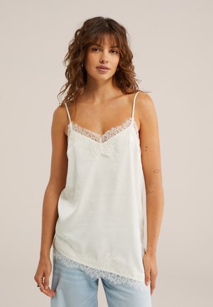 Femme aux cheveux bruns ondulés portant un caraco blanc orné de dentelle et un jean bleu clair, debout devant un fond beige.