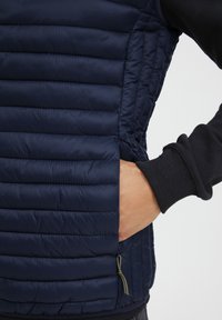 Tamsiai mėlynas pūkinis liemenė su horizontaliais siuvinėjimais, šonine kišene ir elastinėmis juodomis rankogalių detalėmis. Lygi tekstūra su švelniu blizgesiu.