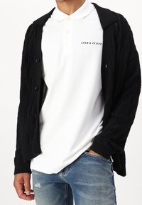 Schwarze Strickjacke mit einem Zopfmuster, getragen über einem weißen Poloshirt mit einem Logo und hellen Knöpfen, kombiniert mit blauen Jeans.