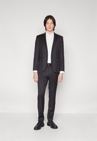 Fato preto com um casaco e calças ajustadas, combinado com uma camisa branca de botões. Apresenta uma textura suave e um design simples. Sapatos pretos completam o visual.