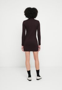 Smaltsittende, langermet kjole i dyp lilla, med høy hals og jevn tekstur. Brukes med knelange svarte sokker og lyse sko.