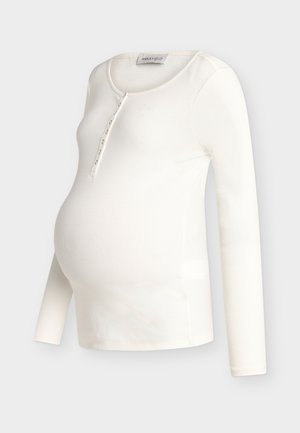 Haut de maternité à manches longues côtelées de couleur crème, avec encolure dégagée et patte de boutonnage, conçu pour s'adapter à un ventre de femme enceinte.