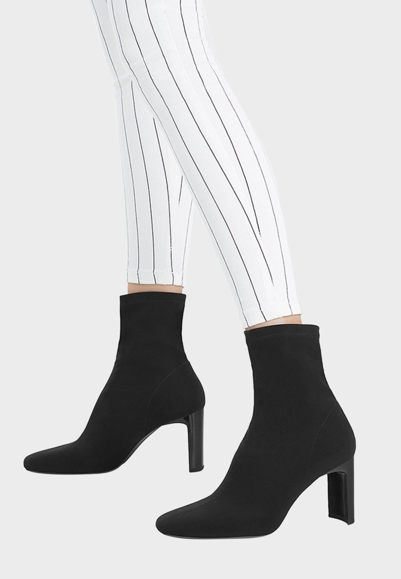 Ben med vita byxor med svarta vertikala ränder, i kombination med svarta klackstövlar som går till mitten av vaden, mot en enhetlig bakgrund.