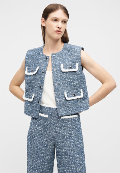 Γυναίκα με μπλε υφασμάτινο γιλέκο χωρίς μανίκια και ταιριαστό παντελόνι με φαρδιά πόδια και ψηλή μέση, με λευκή διακόσμηση, στέκεται με τα χέρια στη μέση.