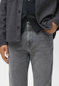 Man in een donkergrijze button-up jas, zwart shirt en vervaagde grijze spijkerbroek, met één hand in de zak en de andere ontspannen aan de zijkant.